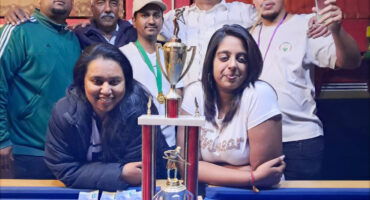 Image of group of people around a pool table with a trophy and cash prizes in the foreground.