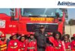 A photo of a group of school children posing with a fire fighter in front of a fire truck.