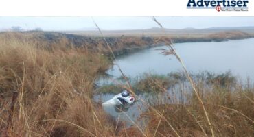 A photo of a white vehicle in a dam surrounded by weeds and grass roots.