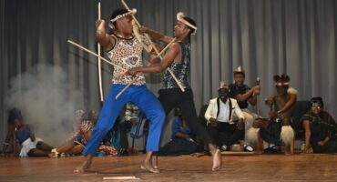 Image of two school learners dressed in traditional Zulu garb stick fighting.