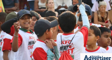 A photo of a group of kids, smiling while wearing white and red KFC Mini Cricket Tshirts and backpacks.