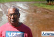 A photo of a muddy road with a man posing in the foreground, wearing a red and blue shirt and glasses.
