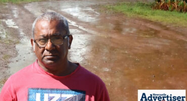 A photo of a muddy road with a man posing in the foreground, wearing a red and blue shirt and glasses.