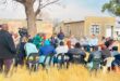 Image of people sitting on chairs in a meeting in a field under a tree.