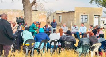Image of people sitting on chairs in a meeting in a field under a tree.
