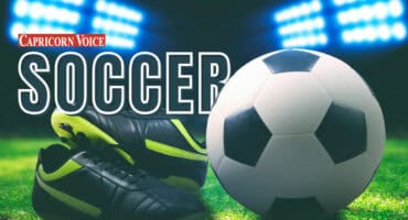 A soccer ball and boots in the foreground with stadium lights in the background.