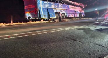 A large, damaged blue and white passenger bus, marked "D&G COACHES," lies on its side off a dark road at night, with its roof partially sheared off and emergency personnel visible nearby.