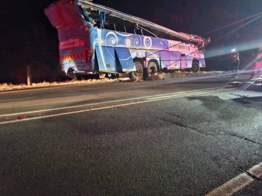 A large, damaged blue and white passenger bus, marked "D&G COACHES," lies on its side off a dark road at night, with its roof partially sheared off and emergency personnel visible nearby.