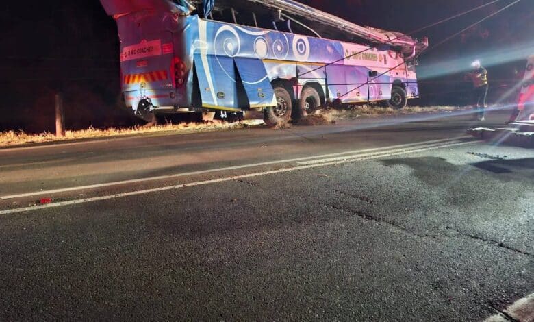 A large, damaged blue and white passenger bus, marked "D&G COACHES," lies on its side off a dark road at night, with its roof partially sheared off and emergency personnel visible nearby.