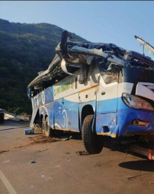 A severely damaged blue and white passenger bus, with its roof mostly destroyed and lying on the roadway on its side after a crash, with a green, mountainous backdrop.
