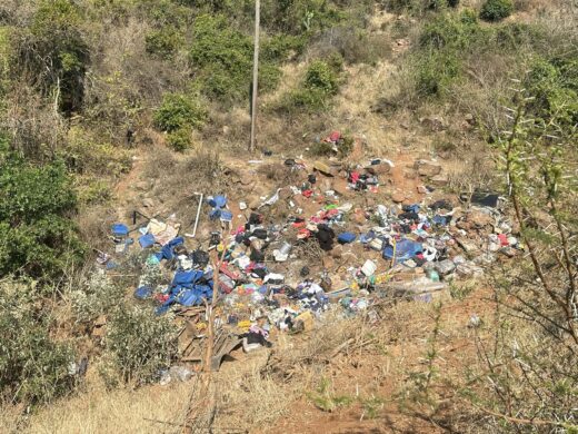 A pile of colorful debris and personal belongings, including blue tarps and clothing, scattered across a steep, dry, scrub-covered embankment beneath a utility pole, suggesting a crash site.