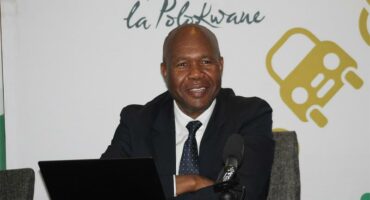 A smiling middle-aged Black man in a dark suit and tie sits at a table with a laptop and microphone in front of a white backdrop that says "la Polokwane" in script and features a yellow car graphic.