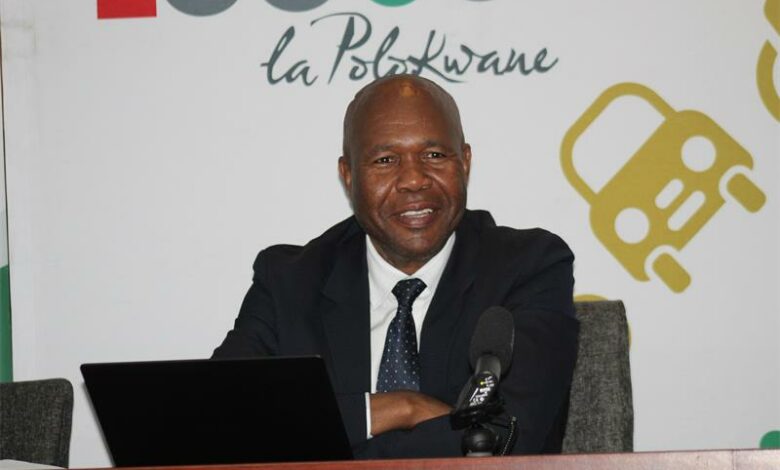A smiling middle-aged Black man in a dark suit and tie sits at a table with a laptop and microphone in front of a white backdrop that says "la Polokwane" in script and features a yellow car graphic.