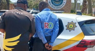 A Hawks officer arrests senior offical outside the Maruleng Local Municipality building, guiding him toward a marked police vehicle parked nearby.