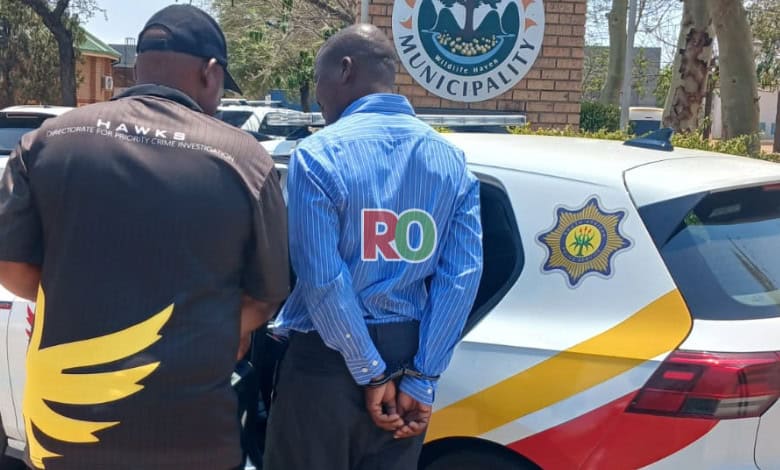 A Hawks officer arrests senior offical outside the Maruleng Local Municipality building, guiding him toward a marked police vehicle parked nearby.