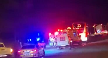 A grainy, low-light night image of an emergency scene on a road showing a police car with flashing blue lights in the foreground, an ambulance, a large white passenger bus, and emergency personnel amidst the red and blue emergency lights.