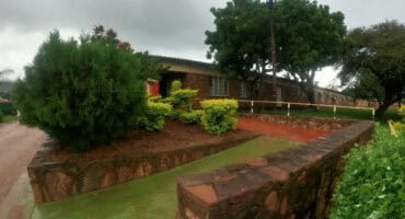 The exterior of a long, single-story brick building at Siloam Hospital, viewed from a landscaped area with a stone retaining wall, hedges, large trees, and a wet, cloudy sky.