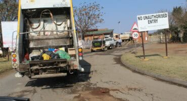 A large white rear-loading garbage truck, with its compactor open and filled with refuse, is backed up onto a dirty paved road at an entrance marked with a "NO UNAUTHORISED ENTRY" sign, leading to a facility under the Polokwane Municipality.