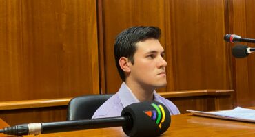 A young adult man with dark hair, wearing a striped collared shirt, sits at a wooden table in a formal setting with wood-paneled walls, looking intently to his right, with a large microphone in the foreground.