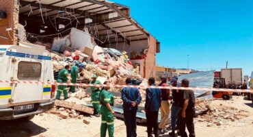 A large pile of rubble and debris spilling from a collapsed red brick building under a bright sky. A police vehicle, workers in green overalls, and a small group of officials stand near a red and white barrier tape marking the wreckage.