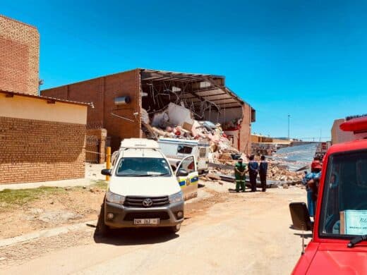 A large section of a single-story brick building has collapsed into a pile of rubble under a bright blue sky, with emergency vehicles (including a white Toyota bakkie and an ambulance) and personnel standing near the site.
