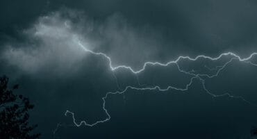 A dramatic photograph captures a brilliant, jagged lightning bolt cracking across a dark, stormy, teal-colored sky at night.
