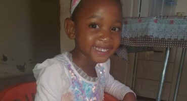 A portrait of a young Black girl smiling brightly while sitting in a small, orange plastic chair indoors. She is wearing a light pink, long-sleeved dress with a sparkly, sequined bodice and a full tulle skirt, and a white and pink patterned headband.