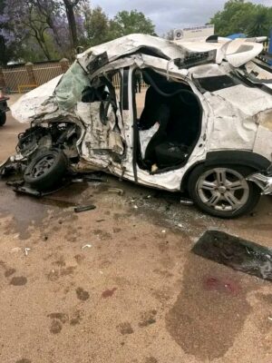 A close-up vertical photo of a severely mangled white passenger car resting on a wet paved surface after a high-impact crash, with its roof, sides, and front end completely crushed and torn apart.