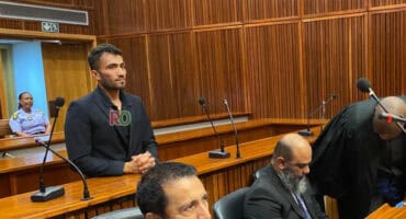 Rameez Patel, a young man in a dark suit, stands in a courtroom dock, looking forward, while lawyers and court officials are seated below him.
