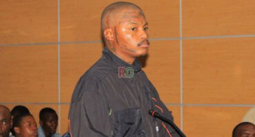 Close-up of Rapper Shebeshxt standing in a courtroom, looking to the side, wearing a black jacket, with visible face and head tattoos.