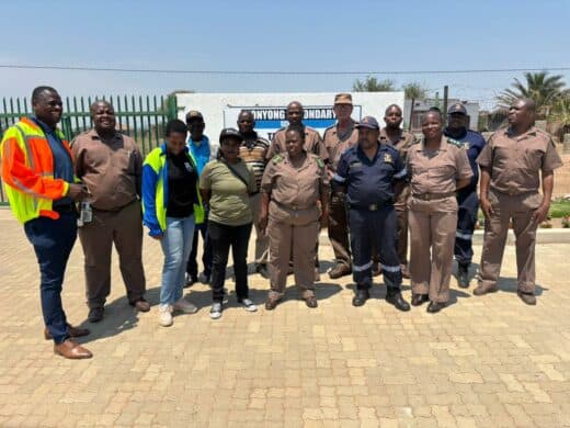 A large group of about 12 individuals, mostly men and women in tan security or uniform attire, posing for a photo outdoors in front of a white wall and a school gate. A sign on the wall is partially visible with the name "NYONG ONDARY" (likely a high school name). The ground is paved with tan bricks, and the sky is bright blue.