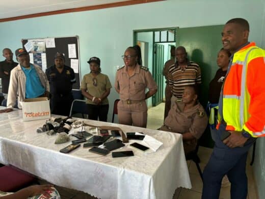 A group of security or law enforcement officials gathered indoors around a table displaying confiscated items, including a box labeled 'Potatrim', numerous cell phones, dark-coloured socks or pouches, and leafy substances (possibly drugs). The officials are mostly in tan uniforms, with one man on the right wearing a bright orange and yellow safety vest.