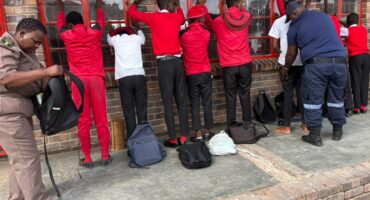 A line of seven male students, most wearing red shirts and black pants, standing with their hands against a red-framed school window while being searched or monitored by officials. Two officials, one in a tan uniform and one in a navy and blue uniform, are present. Backpacks and bags are lined up on the ground in front of the students.