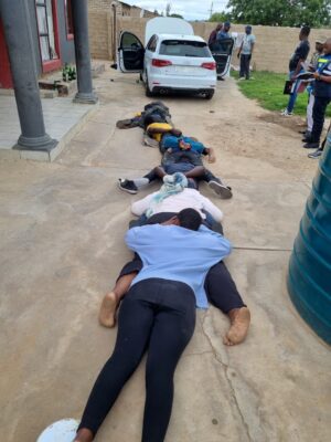 A close-up vertical photograph shows five people lying face down, stacked on top of each other, on a concrete driveway, surrounded by police and onlookers near a white Audi car with open doors.