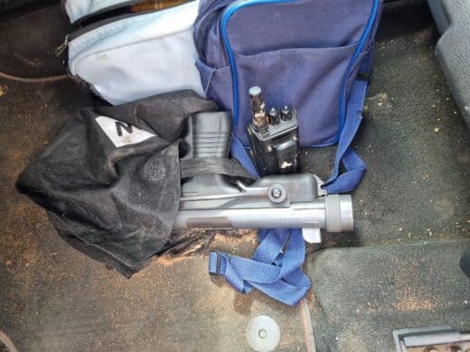A close-up of the dirty floor of a car, showing a disassembled firearm (or non-lethal weapon) partially covered by a black cloth, a walkie-talkie radio, and two blue and light gray bags.