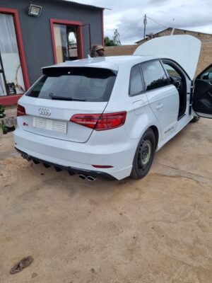 A white Audi hatchback is parked on a dirt/concrete driveway with its bonnet and driver-side rear door open, and a black military-style boot on a person's foot is visible in the background doorway of a dark building. The car has a space-saver spare tire on the rear left wheel.