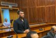 Rameez Patel, a young man in a dark suit, stands in a courtroom dock, looking forward, while lawyers and court officials are seated below him.