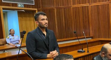 Rameez Patel, a young man in a dark suit, stands in a courtroom dock, looking forward, while lawyers and court officials are seated below him.