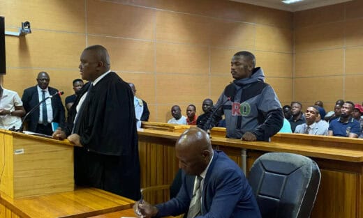 Rapper Shebeshxt standing in a courtroom dock next to his lawyer, who is speaking from a podium.