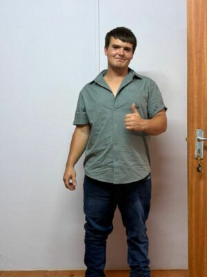 A young man with short brown hair, wearing a short-sleeved, sage-green collared shirt and dark blue jeans, stands indoors against a light-colored wall, giving a thumbs-up with his right hand. A brown wooden door is visible on the right.