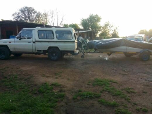 A white, older-model Toyota Land Cruiser 70-series bakkie/truck with a canopy is parked on a dirt patch, towing a trailer covered with a dark tarp, likely carrying a boat. There are trees and structures in the background.
