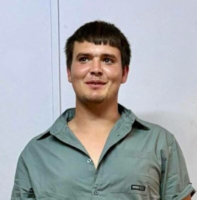 Headshot of a smiling young man with light brown hair cut in short bangs, wearing a light blue-grey collared shirt.