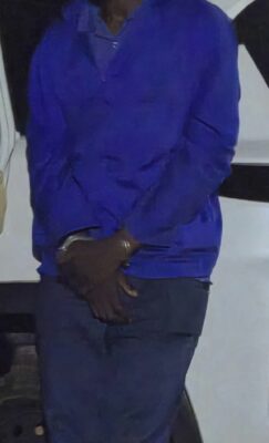 Close-up photograph of an adult person's midsection, wearing a blue jacket and dark pants, with their wrists handcuffed together in front of them.