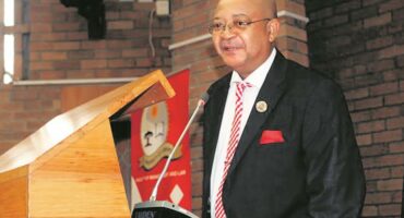 Photograph of Judge President George Phatudi, a bald, light-skinned man wearing glasses, a black pinstripe suit, and a red and white striped tie, standing behind a wooden podium and speaking into a microphone.