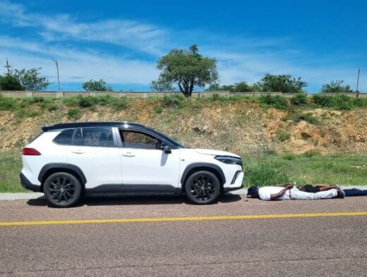 A white Toyota Corolla Cross SUV with a black roof is parked on the shoulder of a highway next to a person lying face-up on the asphalt, positioned near the front bumper.