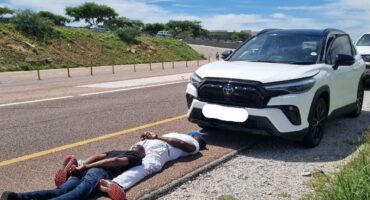 A white Toyota Corolla Cross SUV is parked on a roadside, with two people lying on the ground behind the car's right front tire, facing the camera.