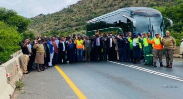 A large group of approximately 30 people, including men and women in various uniforms (police, traffic, and security) and civilian attire, stand on a paved road with their hands raised in a gesture of unity or acknowledgement, in front of a green coach bus.
