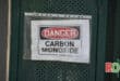 a plastic sleeve with a danger warning placed inside, it says Danger Carbon Monoxide, the plastic sleeve is ziptied to a green metal gate