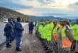 A group of law enforcement and traffic officials are gathered outdoors on a dirt shoulder next to a road, participating in a briefing.