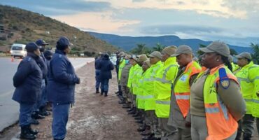 A group of law enforcement and traffic officials are gathered outdoors on a dirt shoulder next to a road, participating in a briefing.